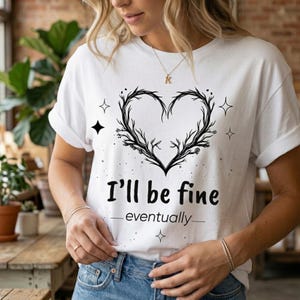 Puede incluir: Camiseta blanca con un diseño negro en forma de corazón hecho de ramas y el texto "I'll be fine - eventually-". La camiseta también presenta pequeñas formas de estrella y diamante.