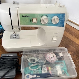Peut inclure: Machine à coudre Brother LS2400 blanche avec pédale et boîte en plastique transparente remplie de fournitures de couture. La machine a un motif floral et des molettes de contrôle. Un livre est visible en arrière-plan.