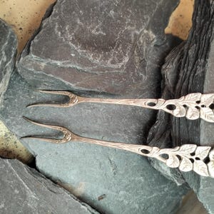 May include: Two silver-toned metal appetizer forks featuring a two-pronged design. The handles are decorated with a detailed relief pattern of leaves and a blooming rose at the end of each handle, with a small rose motif at the base of the tines.