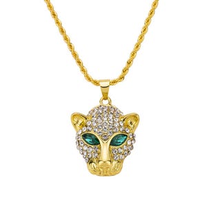Puede incluir: Collar dorado con un colgante con forma de cabeza de leopardo. El colgante está incrustado con cristales transparentes y tiene ojos verde esmeralda. La cadena es de diseño trenzado.