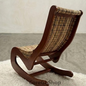 Puede incluir: Sillón balancín de madera con respaldo y asiento curvos. La silla tiene una estructura marrón oscuro y un asiento y respaldo de tela tejida en tonos marrones y beige. La silla está sobre una alfombra blanca.