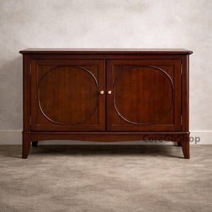 Puede incluir: Un aparador de madera marrón oscuro con dos puertas y una base curva. Cada puerta presenta un diseño circular grande. El mueble tiene pomos dorados y está colocado contra una pared de color neutro.