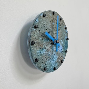 Op de afbeelding: Een ronde wandklok met een getextureerd, gevlekt oppervlak in tinten blauw en grijs. De wijzerplaat heeft zwarte studs die de uren markeren en helderblauwe wijzers. De klok is aan een witte muur bevestigd.