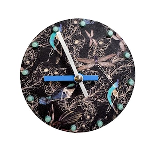 Puede incluir: Reloj de pared redondo con fondo negro con un estampado de libélulas, plumas y pájaros en tonos azules, turquesa y marrón. El reloj tiene un centro plateado, manecillas blancas y un segundero azul. Tachuelas turquesas marcan las horas.