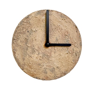 May include: A circular wall clock with a textured, sandy-coloured face. The black clock hands indicate approximately 3 o'clock. The clock has a rustic, earthy aesthetic, suitable for home decor.