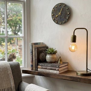 Puede incluir: Un reloj de pared redondo con esfera azul grisáceo y agujas doradas cuelga de la pared. Una estantería de madera debajo contiene libros, una pequeña planta y una lámpara de latón. Un sillón gris con una manta beige está en primer plano.