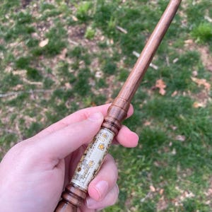 May include: A wooden wand with a brown and white design. The wand has a pointed tip and a decorative handle. The handle is wrapped in a white material with a gold pattern.