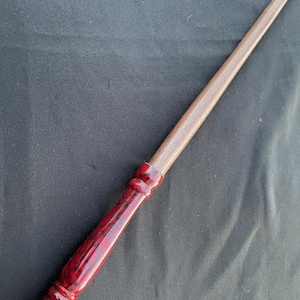 May include: A handcrafted wooden wand with a dark red handle and a light brown shaft. The handle has a glossy finish with black marbling. The wand's tip is pointed. The wand is a magical item.