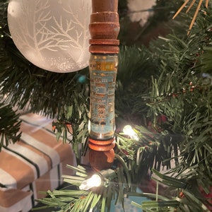 May include: A unique Christmas tree ornament with a wooden stem and a cylindrical glass section featuring a circuit board design. The ornament is adorned with a light blue and gold pattern, and is hanging on a green Christmas tree with white lights.