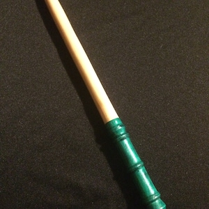 Può includere: Una bacchetta di legno con una punta dipinta di verde. La bacchetta è lunga circa 30 cm e ha una finitura liscia e naturale.