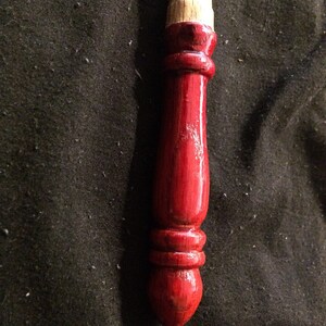 May include: A close-up of a red wooden spindle with a natural wood top. The spindle is smooth and polished, with a rounded tip.