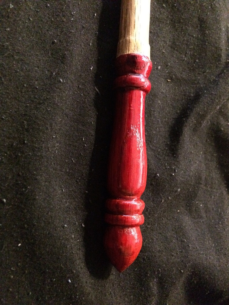 May include: A close-up of a red wooden spindle with a natural wood top. The spindle is smooth and polished, with a rounded tip.