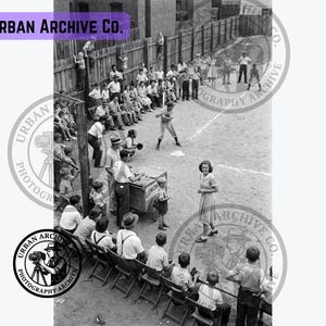 Può includere: Una fotografia in bianco e nero che mostra una partita di baseball con un battitore alla casa base, spettatori seduti su sedie pieghevoli e un piccolo carretto con l'etichetta LEMONADE. La scena include una recinzione in legno e numerose persone.