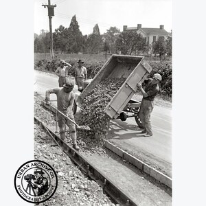 May include: A black and white historical photograph showing four workers constructing a road. One worker uses a shovel to spread gravel while a dump trailer is tilted to release a load of stones. A circular logo with the text URBAN ARCHIVE CO. PHOTOGRAPHY-ARCHIVE is in the corner.