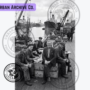 May include: A black and white historical photograph showing a group of dockworkers sitting on wooden crates at a harbor. In the background, large cargo ships are docked, and a horse-drawn cart is visible. The text Urban Archive Co. appears.