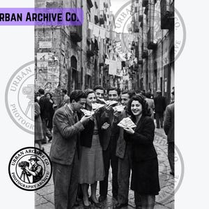 Op de afbeelding: Een zwart-witfoto getiteld Urban Archive Co. met een groep mensen die in een smalle stadsstraat staan en stukken pizza eten. De personen dragen kleding uit het midden van de twintigste eeuw, met wasgoed dat boven de straat hangt te drogen.