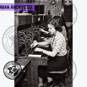 May include: A black and white photograph showing a telephone operator wearing a headset, seated at a large manual switchboard with numerous cords and plugs. The operator is actively working at the console, with another operator visible in the background.