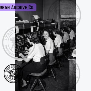 May include: A black and white photograph showing a row of operators at a telephone switchboard. Each operator wears a headset and sits on a swivel chair. One operator looks toward the camera. The text Urban Archive Co. appears in the image.