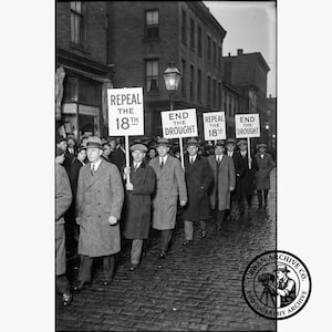 May include: A black and white historical photograph showing a group of people walking down a cobblestone street carrying signs that read REPEAL THE 18TH and END THE DROUGHT. The individuals are wearing long coats and hats in an urban setting.