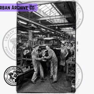 Peut inclure: Une photographie en noir et blanc intitulée Urban Archive Co. montrant une chaîne de montage automobile de 1940. Des ouvriers en salopettes et chapeaux sont réunis autour d'un châssis de voiture sombre dans une grande usine industrielle.