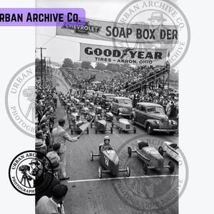 May include: A black and white photograph of a Soap Box Derby race with small gravity-powered cars on a track. Banners for Chevrolet and Goodyear Tires are visible overhead. A person waves a checkered flag as the cars approach the finish line.