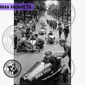 May include: A black and white photograph of a soapbox derby race on a paved street. A child sits in a small, custom-built car labeled Akron Special in the foreground. Other children and adults are visible in the background with additional race cars.
