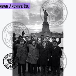 May include: A black and white photograph featuring a group of children waving in front of the Statue of Liberty. The text Urban Archive Co. appears in the top left corner and within circular watermarks overlaid on the image.