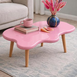 May include: A pink, kidney-shaped coffee table with light wood legs. A blue vase with pink flowers, a pink mug, and a book sit on the table. Decorative seashells add a coastal touch to the living room decor.