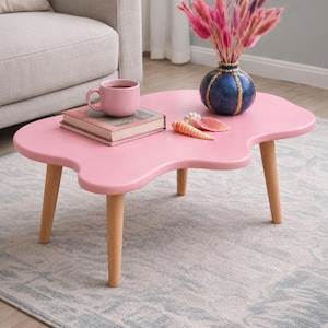 May include: A pink, kidney-shaped coffee table with light wood legs. The table is decorated with a pink mug, a book, a blue vase with pink flowers, and seashells. The table is set on a patterned rug.
