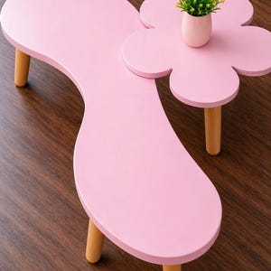 May include: A pink, flower-shaped coffee table with three wooden legs. A smaller, matching table with a flower shape is attached. A small pink pot with a green plant sits on top of the smaller table.