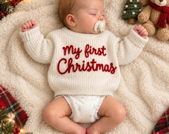 Op de afbeelding: Een slapende baby draagt een crèmekleurige gebreide trui met lange mouwen en de rode geborduurde tekst My first Christmas op de voorkant. De baby heeft een witte speen in de mond en draagt een witte luier met een klein boompjespatroon.