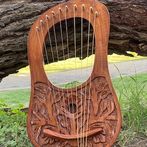 May include: A wooden lyre, featuring carved floral and tree patterns. The instrument has a curved body, a sound hole, and multiple strings. The lyre is a warm brown colour and rests on green grass. The lyre is approximately 60 cm in height.