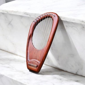 May include: A wooden lyre in a deep reddish-brown hue. The instrument has a curved, open frame with multiple silver strings stretched across. It rests against a white marble surface, highlighting its classic design. The lyre's dimensions are approximately 50 cm in height.
