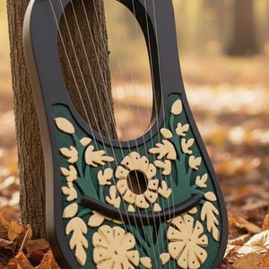 May include: A black lyre with a green and cream floral design, leaning against a tree. The instrument has strings and is decorated with cut-out flowers and leaves. The background is blurred with fall foliage.