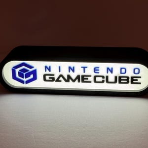 May include: A rectangular, black-framed, illuminated sign with rounded edges featuring the blue Nintendo GameCube logo on the left and the text NINTENDO GAMECUBE in black lettering on a white, backlit background, emitting a soft glow.