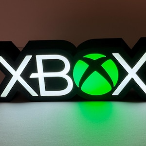 Puede incluir: Un letrero negro tridimensional que muestra la palabra XBOX en una fuente negrita y cuadrada. Las letras X, B y X están iluminadas con una luz blanca brillante, mientras que la O circular presenta un logotipo de esfera Xbox verde vibrante.