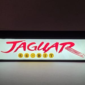 May include: A rectangular, illuminated sign featuring the red text JAGUAR in a stylized font, with the yellow text 6-4-BIT positioned below it inside yellow circles. The sign has a black frame and emits a bright white light from the center panel.