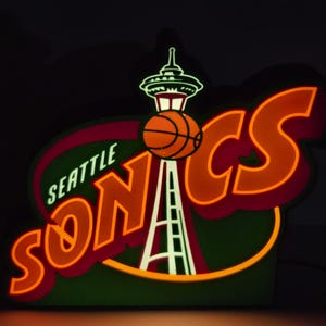 Puede incluir: Un letrero luminoso de estilo neón con el texto SEATTLE SONICS en letras naranjas. El diseño incorpora un gráfico de la Space Needle en blanco y verde, con un balón de baloncesto naranja posicionado frente a la estructura de la torre.