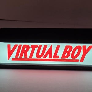 Könnte beinhalten: Ein rechteckiger Leuchtkasten mit schwarzem Rahmen und abgerundeten Ecken, der den Text VIRTUAL BOY in fetten, roten Großbuchstaben auf einem weißen, beleuchteten Hintergrund zeigt. Das Schild ist horizontal ausgerichtet und leuchtet hell.