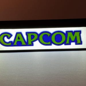 May include: A rectangular, illuminated sign featuring the word CAPCOM in bold, lime green letters with a dark blue outline. The text is set against a bright white, glowing background, all encased within a solid black frame.