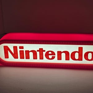 Pode incluir: Um letreiro retangular da Nintendo em forma de pílula com bordas arredondadas. O letreiro apresenta um painel central branco com a palavra Nintendo impressa em letras vermelhas em negrito. A moldura externa brilha com uma luz vermelha.