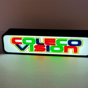 Peut inclure: Une enseigne LED rectangulaire à cadre noir affichant le texte lumineux "COLECO VISION" dans une police grasse sans empattement multicolore. Les lettres sont en rouge, bleu et vert sur un fond blanc brillant et lumineux.