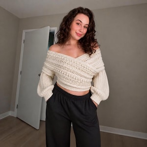 Puede incluir: Un suéter color crema de hombros descubiertos y manga larga con un patrón de punto texturizado, filas horizontales de pompones y un diseño cruzado en la parte delantera, combinado con pantalones negros de tiro alto, corte holgado y cintura elástica.