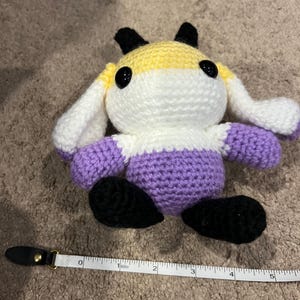 Peut inclure: Une peluche crochet&eacute;e avec une partie sup&eacute;rieure jaune, une partie centrale blanche et une partie inf&eacute;rieure violette, dot&eacute;e d'oreilles et de pieds noirs. Elle a deux grands yeux en plastique noir. Un ruban &agrave; mesurer indique 12,7 cm de large.