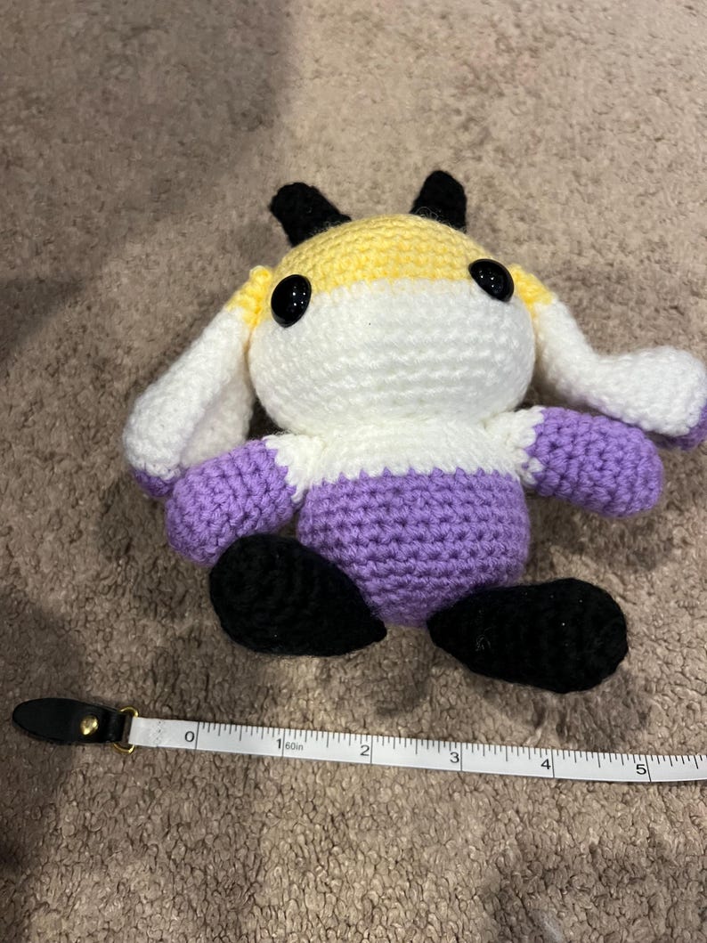 Peut inclure: Une peluche crochet&eacute;e avec une partie sup&eacute;rieure jaune, une partie centrale blanche et une partie inf&eacute;rieure violette, dot&eacute;e d'oreilles et de pieds noirs. Elle a deux grands yeux en plastique noir. Un ruban &agrave; mesurer indique 12,7 cm de large.