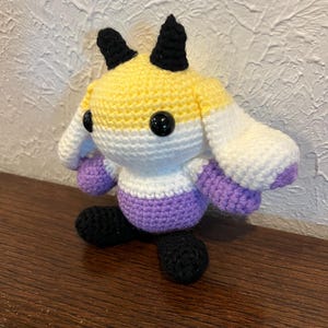 May include: A crocheted plush toy featuring a round head with yellow, white, and purple horizontal stripes, two black horns, and black safety eyes. The body is purple with black feet and long, floppy white ears with purple tips on the sides.