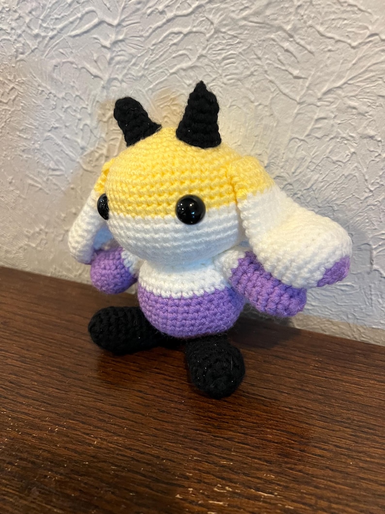 Peut inclure: Une peluche crochet&eacute;e avec une t&ecirc;te ronde &agrave; rayures horizontales jaunes, blanches et violettes, deux cornes noires et des yeux de s&eacute;curit&eacute; noirs. Le corps est violet avec des pieds noirs et de longues oreilles blanches tombantes aux pointes violettes.