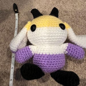 Peut inclure: Une peluche crochet&eacute;e avec un corps jaune, blanc et violet, des oreilles et des pieds noirs, de grands yeux de s&eacute;curit&eacute; noirs, et un ruban &agrave; mesurer blanc plac&eacute; &agrave; gauche indiquant une hauteur d'environ 18 cm de haut en bas.