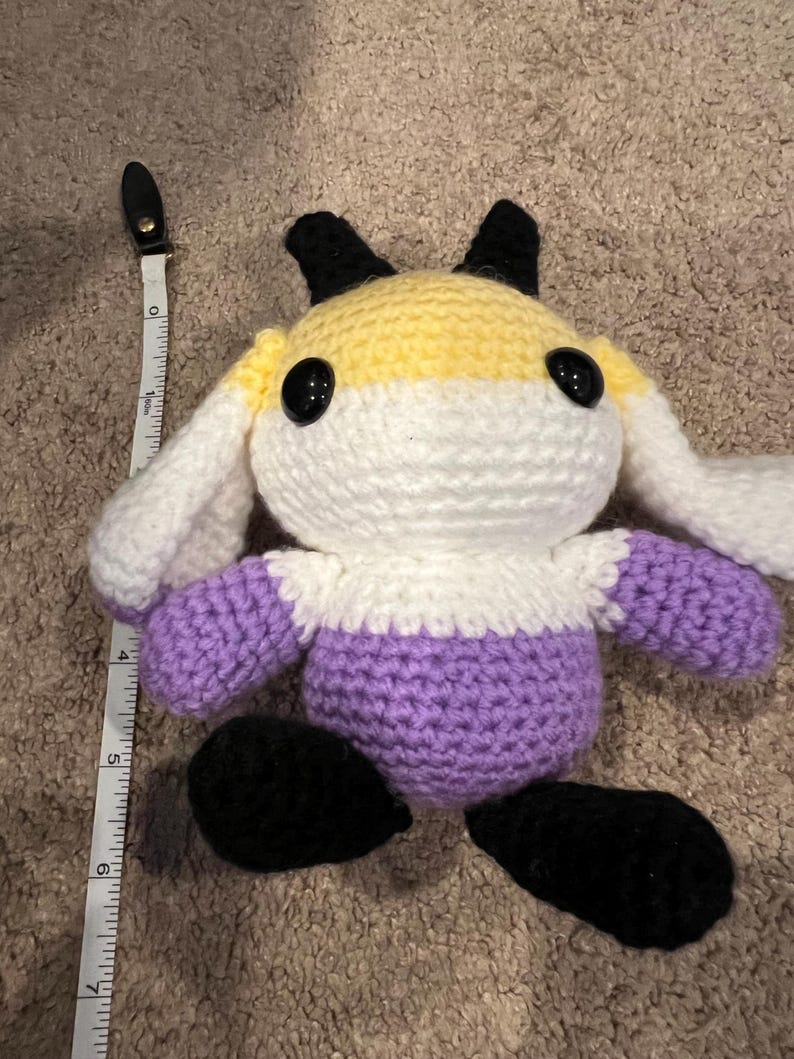 Peut inclure: Une peluche crochet&eacute;e avec un corps jaune, blanc et violet, des oreilles et des pieds noirs, de grands yeux de s&eacute;curit&eacute; noirs, et un ruban &agrave; mesurer blanc plac&eacute; &agrave; gauche indiquant une hauteur d'environ 18 cm de haut en bas.