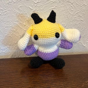 Peut inclure: Une peluche crochet&eacute;e &agrave; la main avec une t&ecirc;te ronde &agrave; rayures horizontales jaunes, blanches et violettes, deux cornes noires et deux yeux en plastique noir. Le corps est blanc et violet avec des pieds noirs et de longues oreilles blanches tombantes.